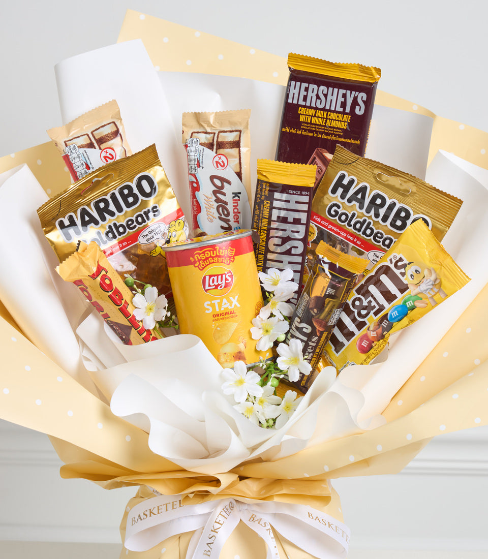 A decorative gift bouquet with yellow and white wrapping containing assorted snacks including Haribo, Hershey's, Lay's, and M&M's, adorned with white flowers and a cream ribbon bow.