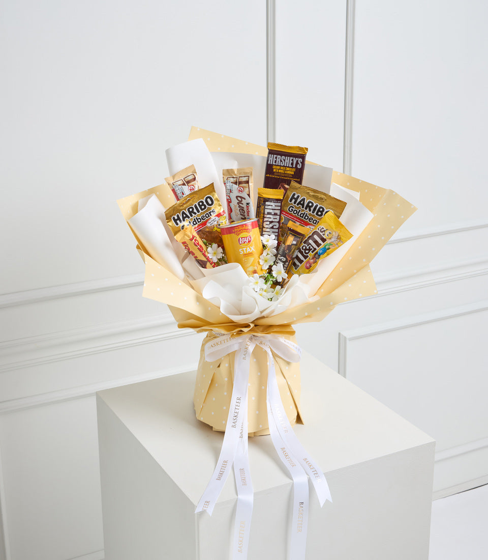 Gold and white snack bouquet with candy, chips, and chocolate arranged in polka dot paper with white ribbon bow on white pedestal.