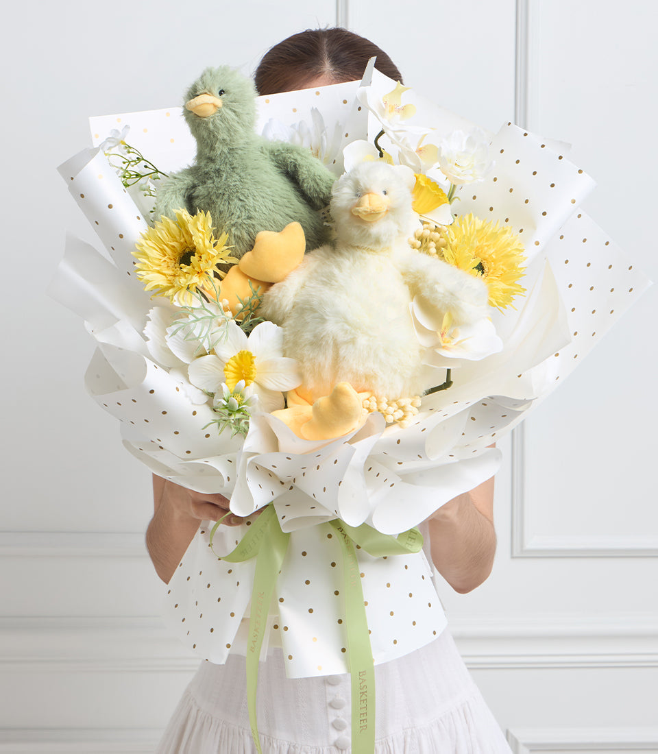 Person holding luxurious bouquet with two plush chick toys, yellow and white flowers, cream wrapping with gold polka dots and green ribbon.