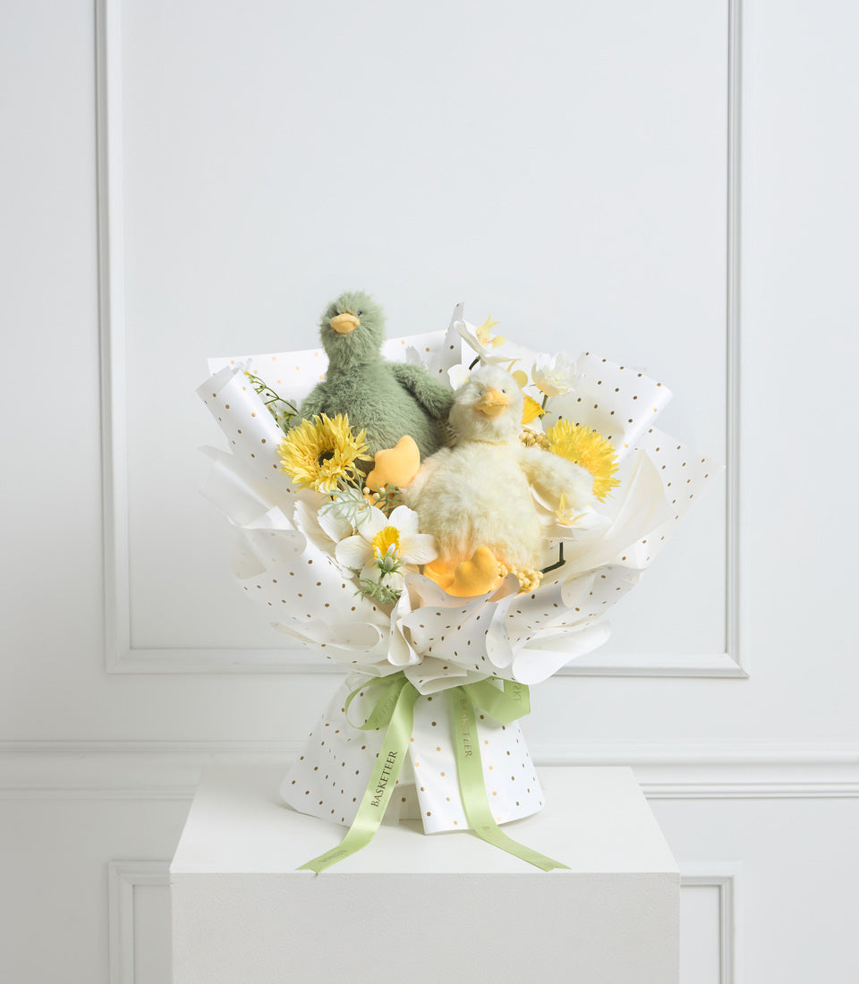 Whimsical gift bouquet with plush bird toys, yellow gerberas, white flowers, and green ribbon on white polka-dot wrapped base.