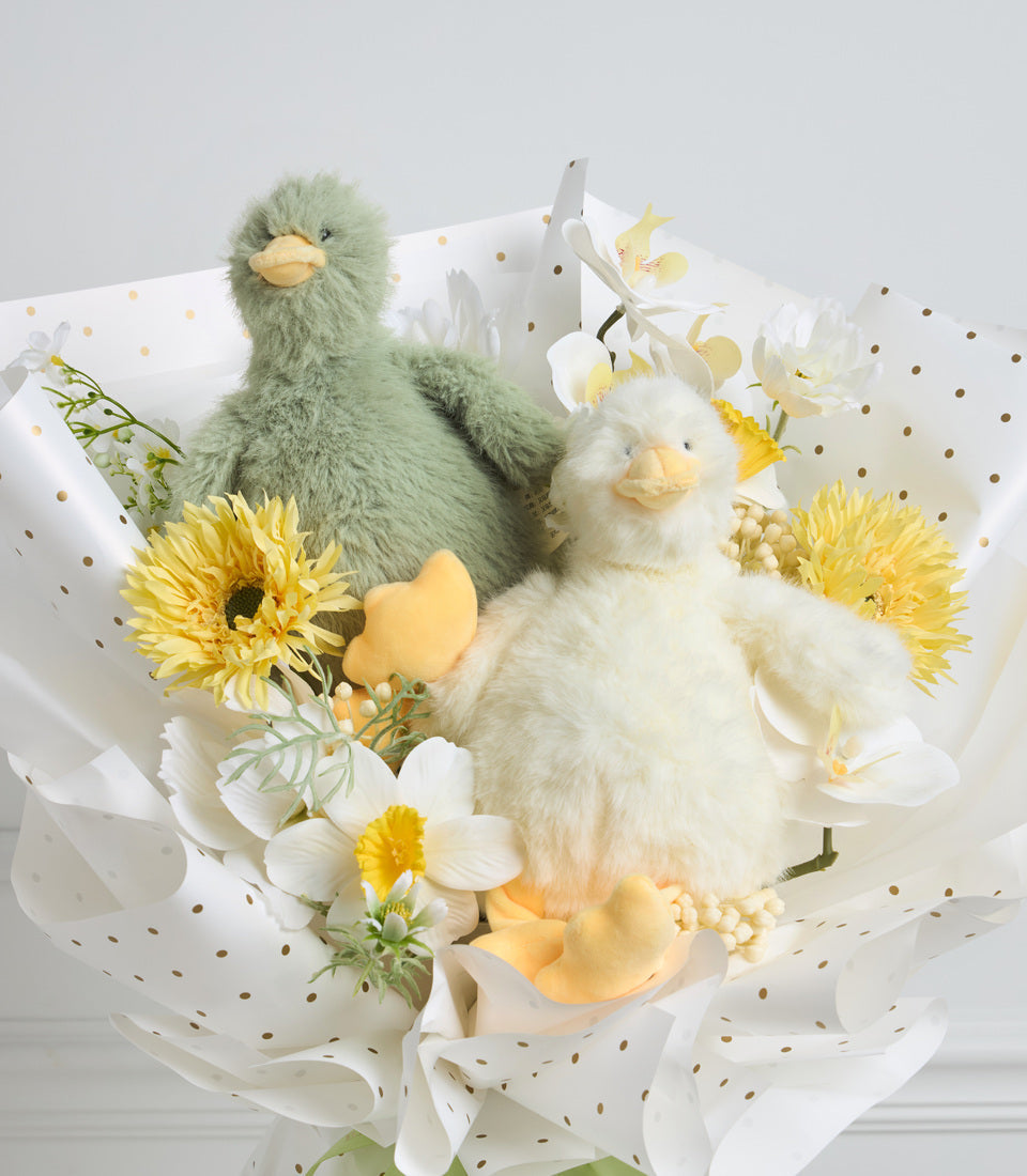 # Alt Text

Fluffy plush chick figurines in sage green and cream with yellow beaks, surrounded by fresh yellow gerberas, white orchids, and pearl accents in white spotted wrapping.