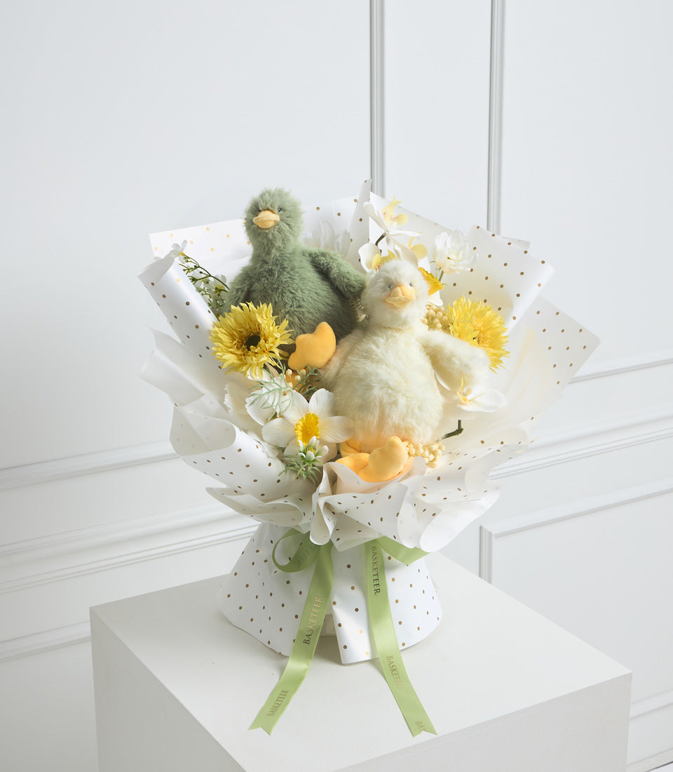 Plush bird bouquet with yellow sunflowers and white flowers, wrapped in polka dot paper with green ribbon, displayed on white pedestal.